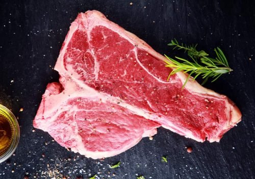 Uncooked t-bone or porterhouse steak seasoned with salt and spices with fresh herbs with a glass of whiskey above